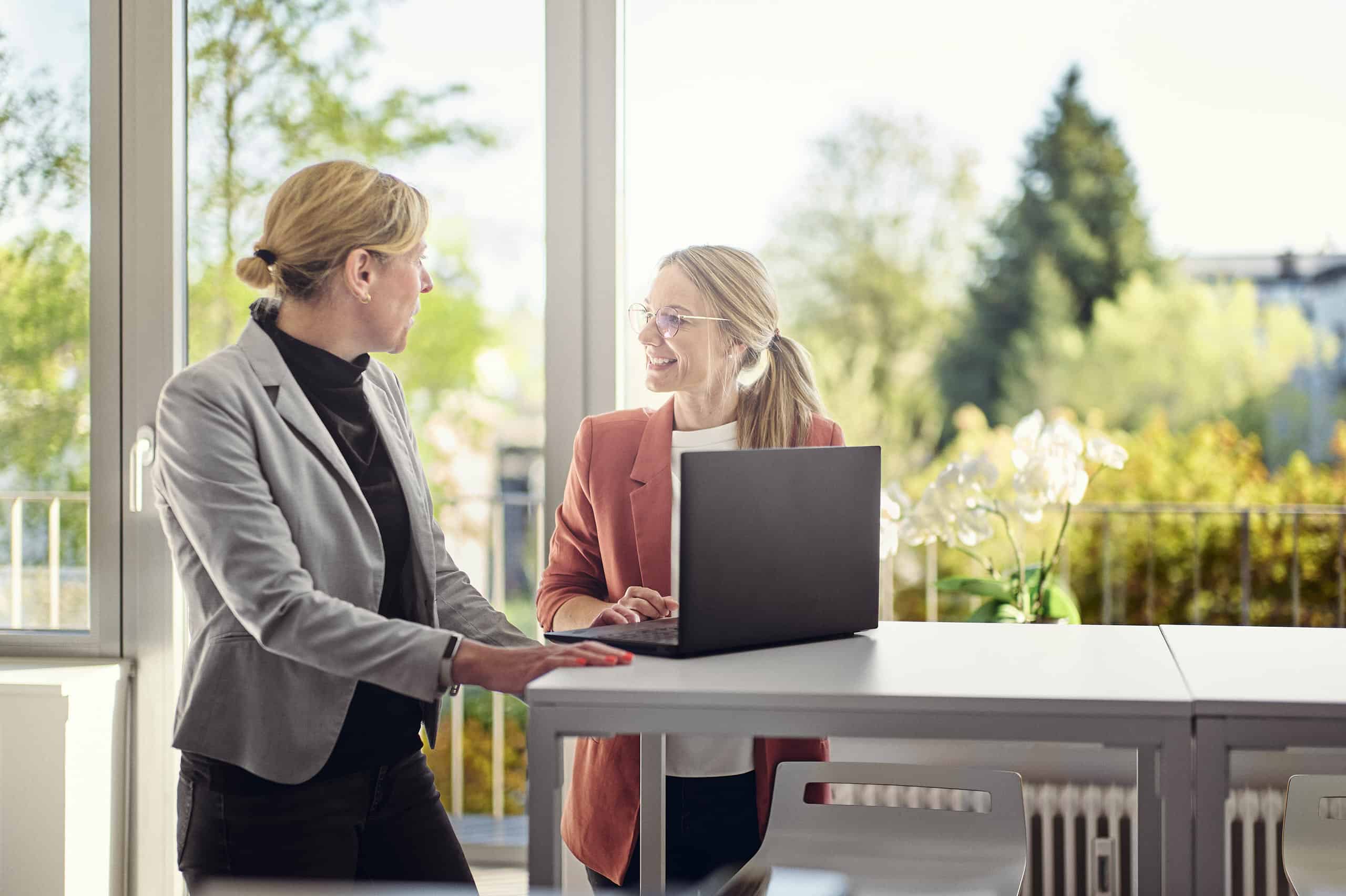 Celine Spitz i Magdalena Sabaß stoją w salonie przy stoliku barowym, na którym leży laptop, i rozmawiają ze sobą.