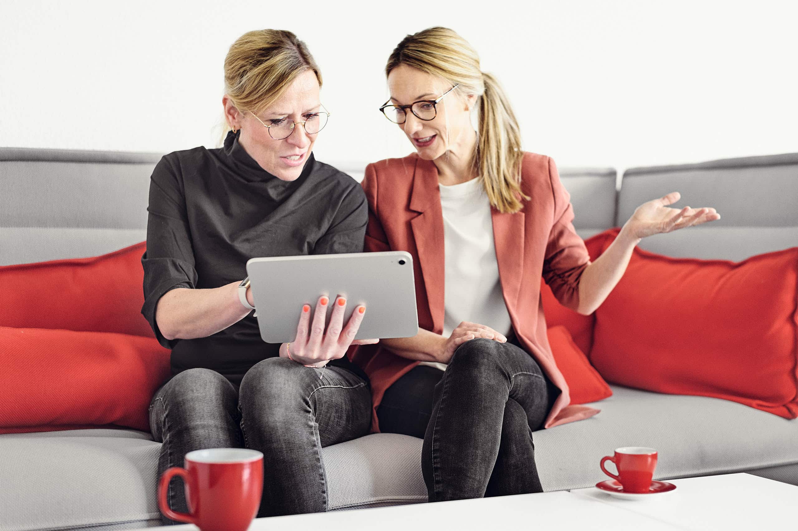 Magdalena Sabaß explains something to Celine Spitz on a tablet while they sit on a couch with coffee on the coffee table.