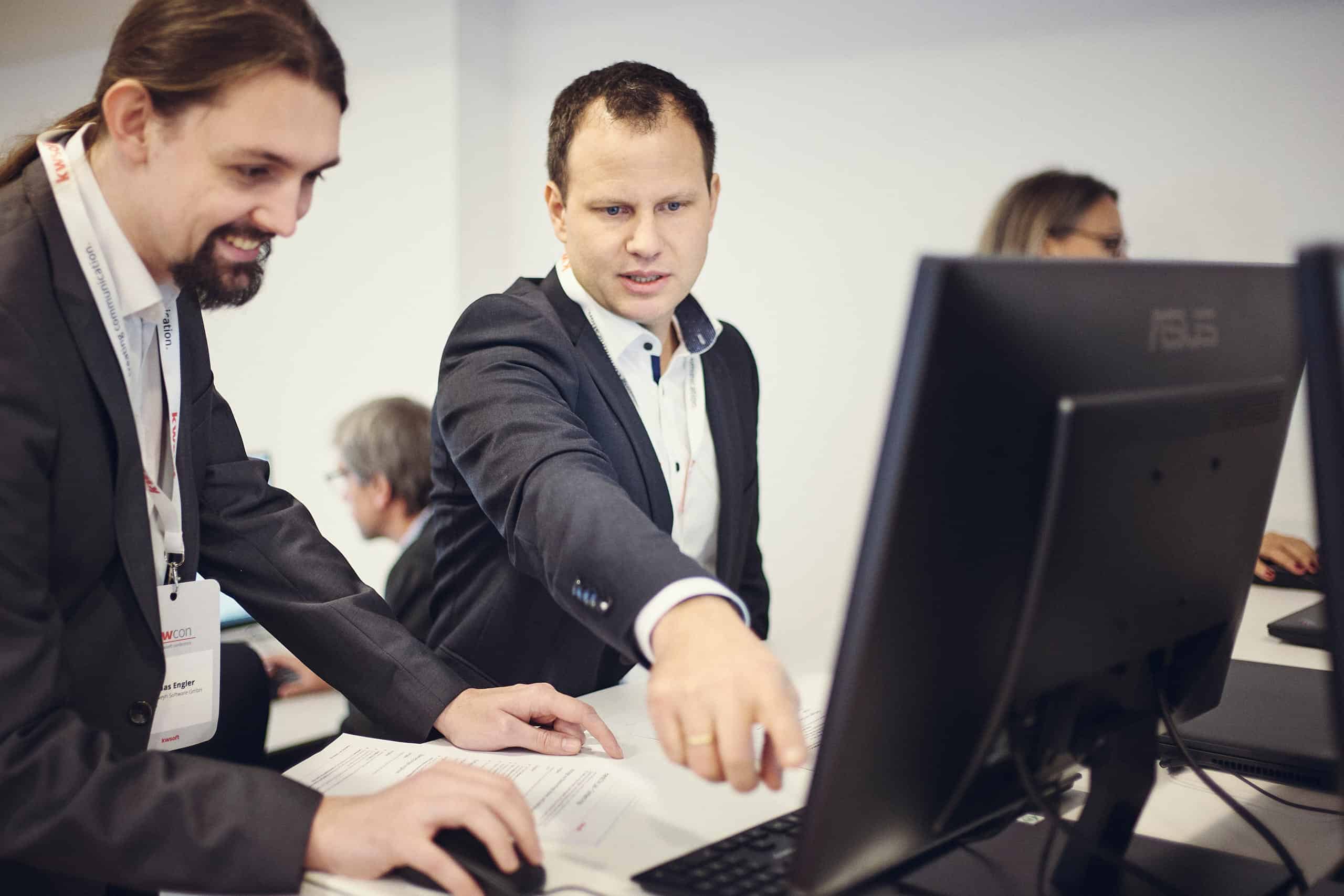Two men in suits are looking at a screen, with the man on the right pointing at the screen and the other operating the mouse..
